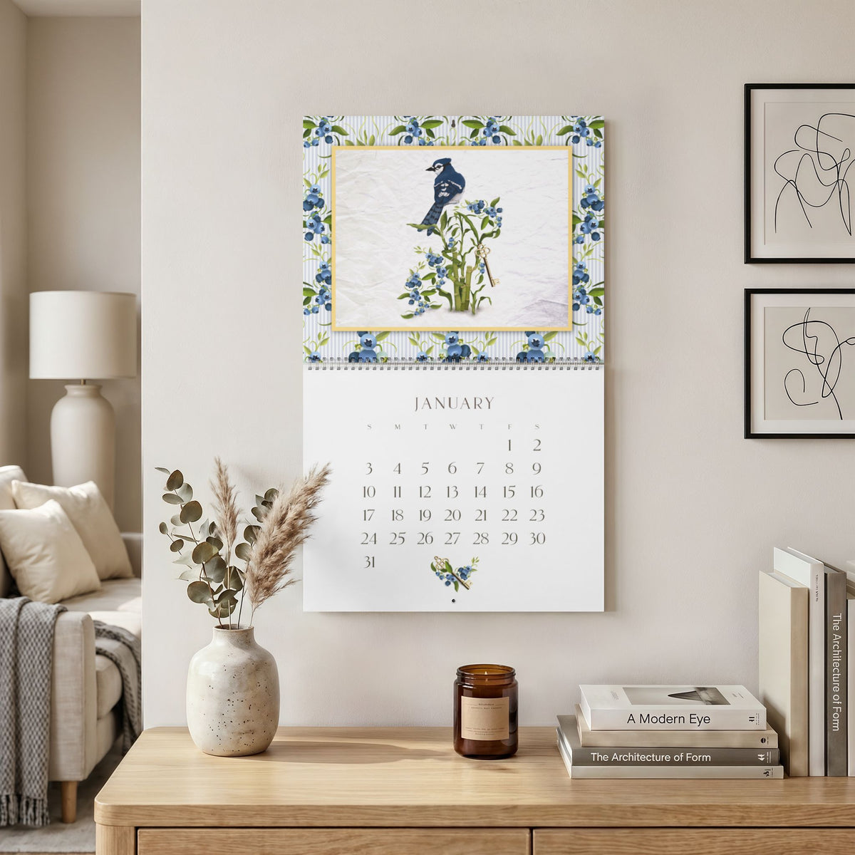 2027 Sue Krizman wall calendar hanging in a neutral home setting, showing the January Blue Jay illustration
