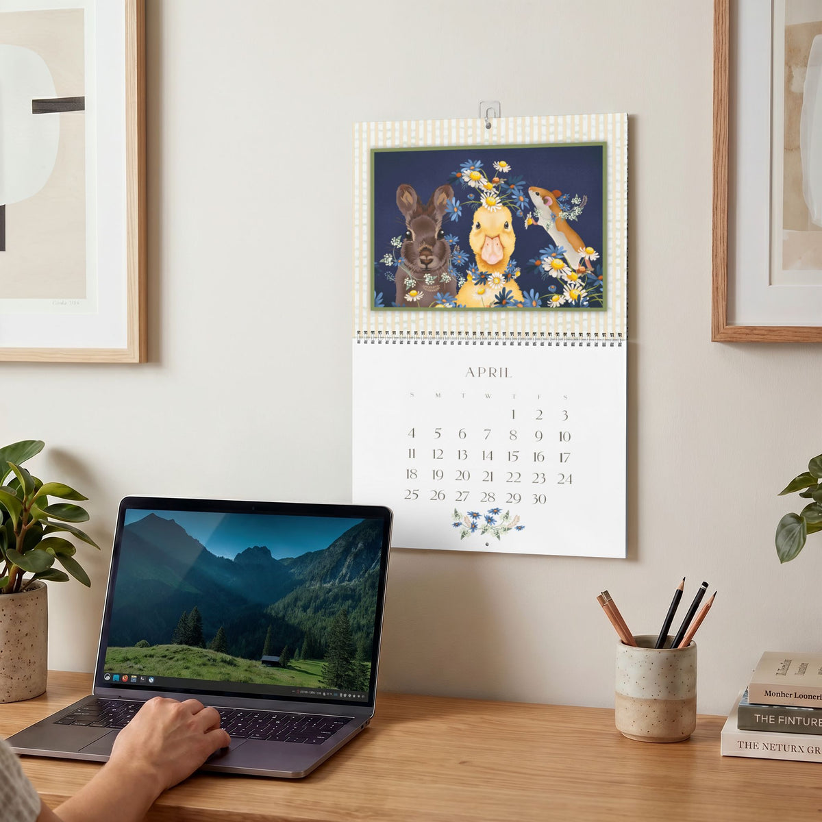 2027 Sue Krizman wall calendar hanging above a desk, featuring the April illustration of colorful animals and flowers