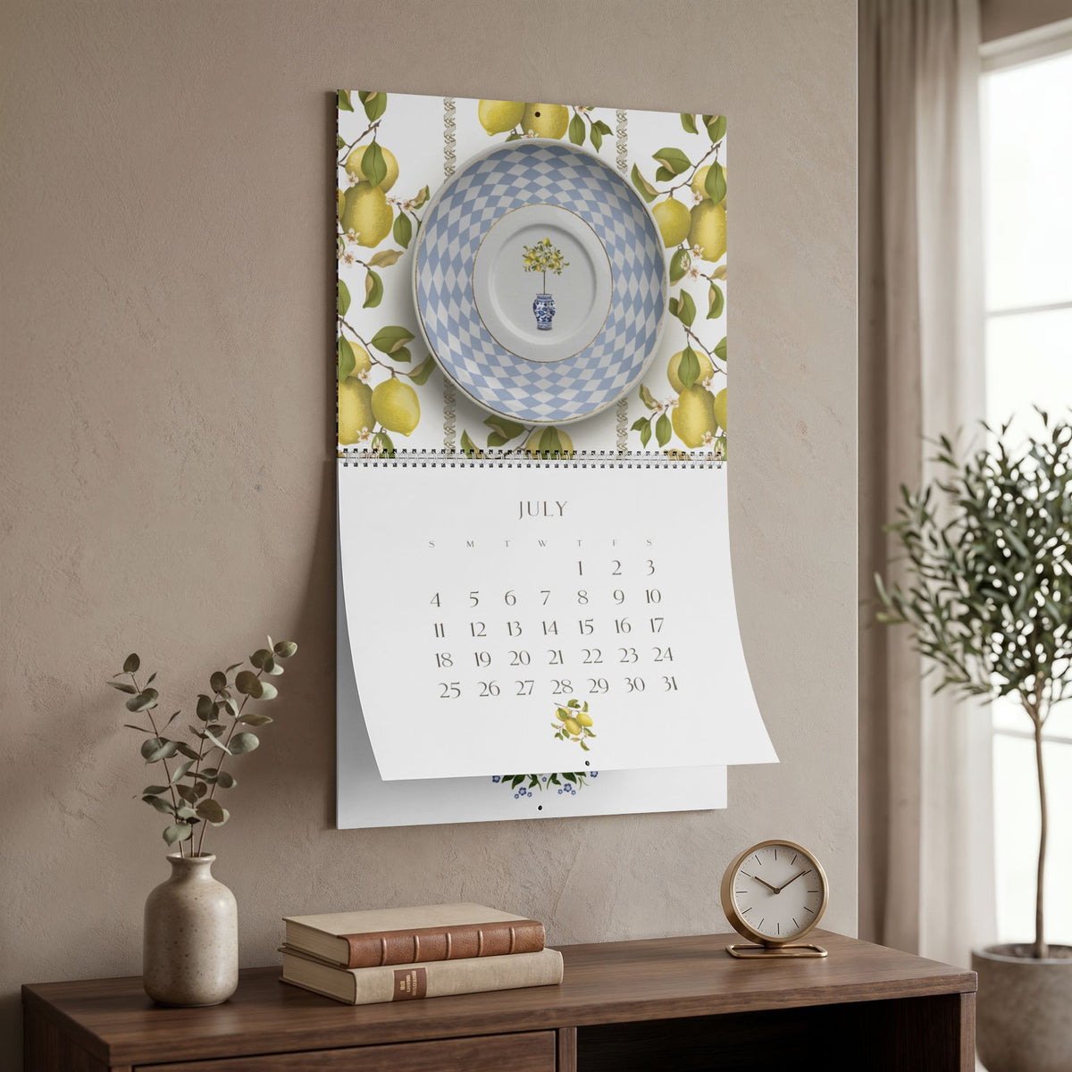 2027 Sue Krizman wall calendar hanging in a home setting, featuring the July illustration with lemons and a blue patterned plate